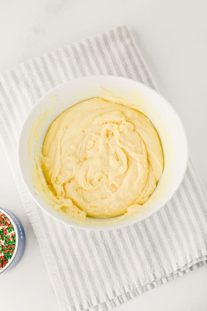 Overhead view of a bowl of melted white chocolate condensed milk fudge mixture, with a small bowl of holiday sprinkles beside it.