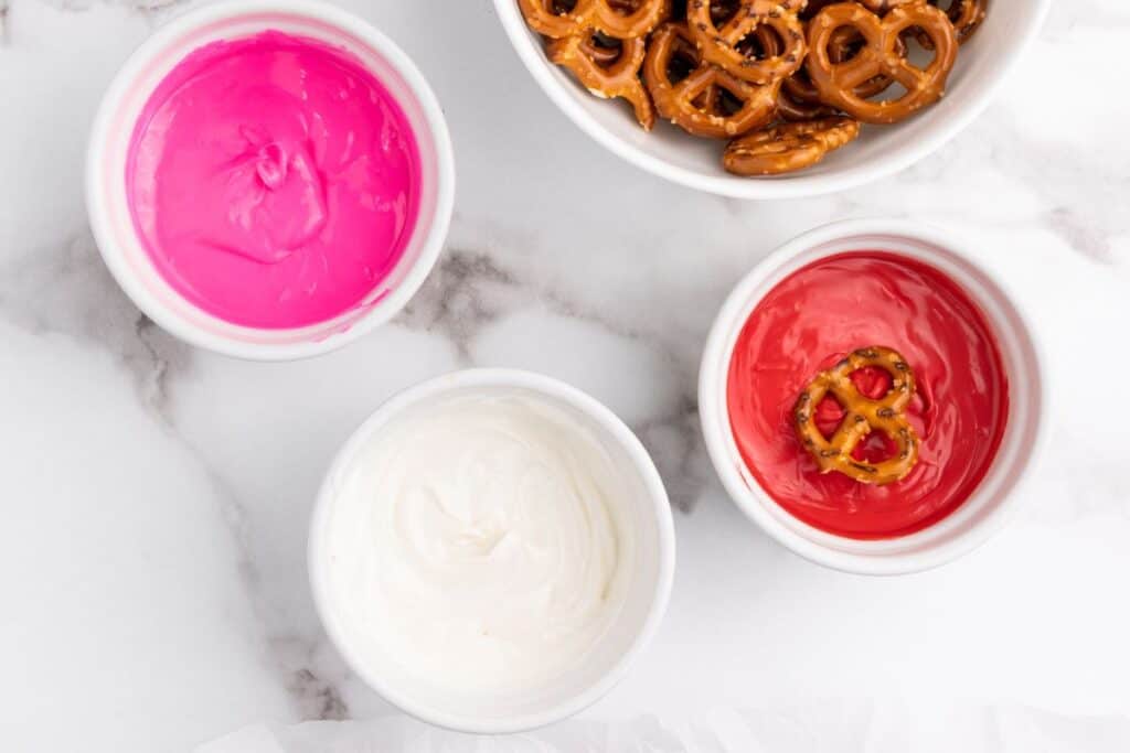 Small bowls of red, pink and white melted candy melts, with one mini pretzel dipped in the red candy melt bowl.