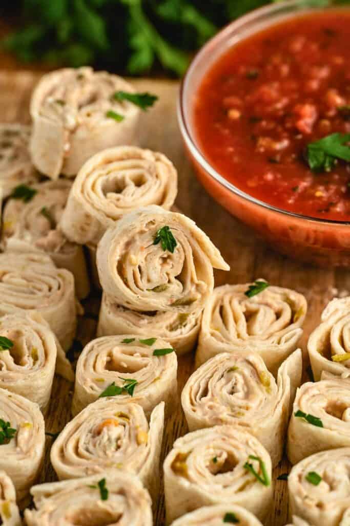 Sliced Mexican burrito roll ups arranged on a wooden board with visible layers of tortilla and creamy filling dotted with herbs and small vegetable pieces. A bowl of red salsa sits beside them making this a great travel snack idea for dipping and easy serving.