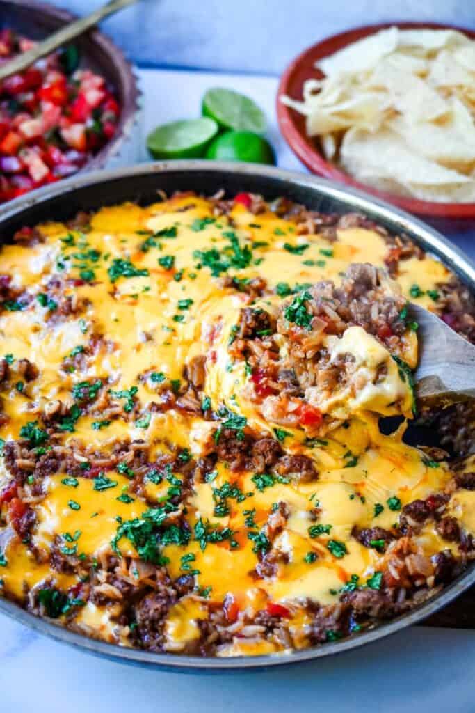 Cheesy taco skillet with ground beef and rice topped with melted cheddar and chopped cilantro as a spoon lifts a scoop from the pan. Bowls of tortilla chips, fresh pico de gallo, and halved limes sit in the background showing an easy 30 minute meal served straight from the skillet.
