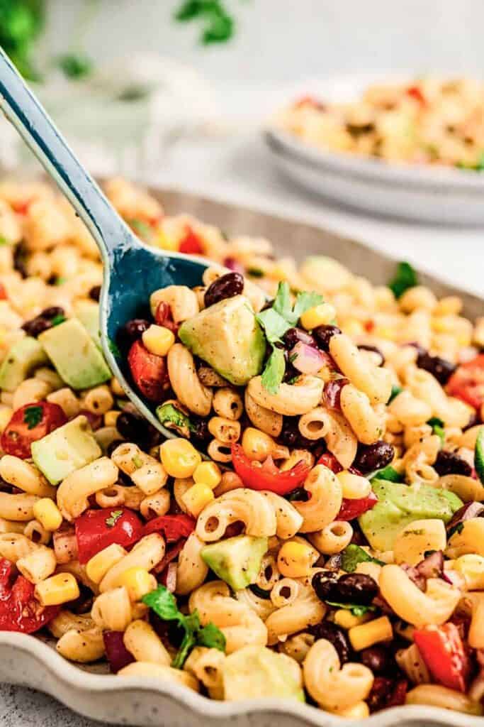 Colorful Mexican macaroni salad served in a large dish, featuring elbow pasta tossed with black beans, corn, avocado chunks, cherry tomatoes, red onion, and fresh cilantro. A blue serving spoon lifts a generous scoop of the zesty, seasoned salad, perfect for a vibrant side dish.