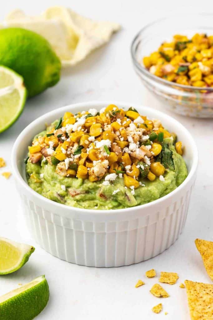 Mexican Street Corn Guacamole served in a white ramekin, topped with charred corn, crumbled cotija cheese, chopped onions, and fresh herbs. Surrounded by lime wedges and tortilla chips, this vibrant dip blends creamy avocado with bold, zesty street corn flavors.