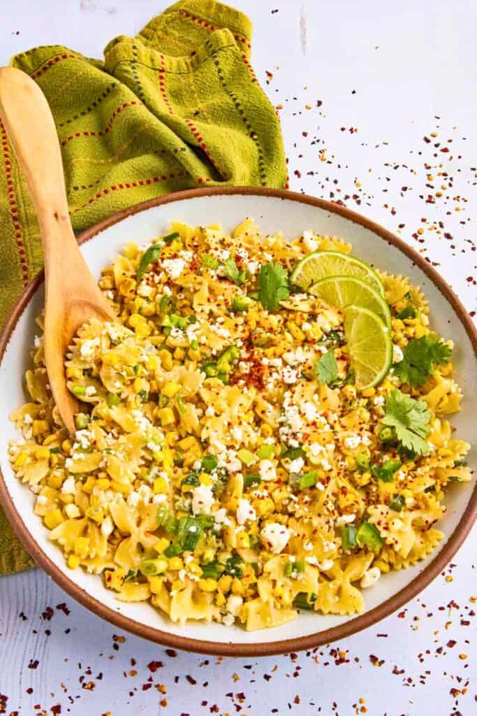 Mexican street corn pasta salad served in a large ceramic bowl, featuring bowtie pasta mixed with roasted corn, crumbled cotija cheese, chopped green onions, cilantro, and a sprinkle of chili flakes. Garnished with lime slices and accompanied by a wooden serving spoon and a green cloth napkin.
