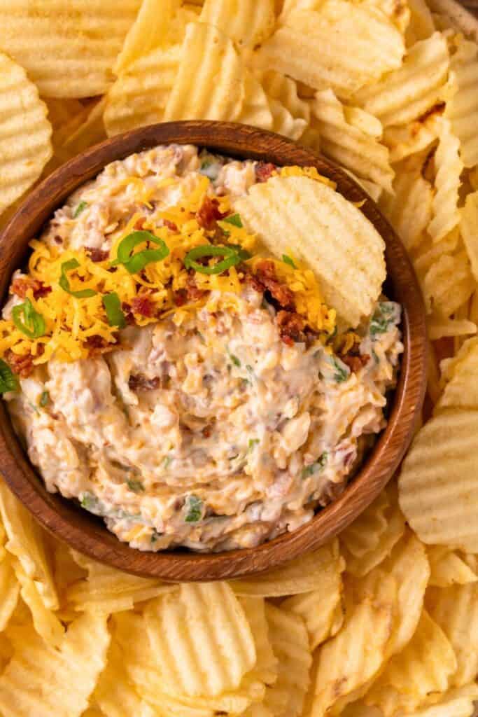 A wooden bowl filled with creamy million dollar dip, topped with shredded cheddar cheese, crispy bacon bits, and sliced green onions, surrounded by a generous serving of ridged potato chips for dipping.