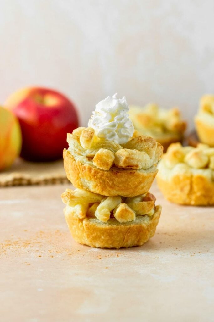 Two mini apple pies are stacked on top of each other, with the top pie topped with a swirl of whipped cream. The golden lattice crust and diced apple filling make these bite-sized treats a charming and festive option for a Thanksgiving dessert.