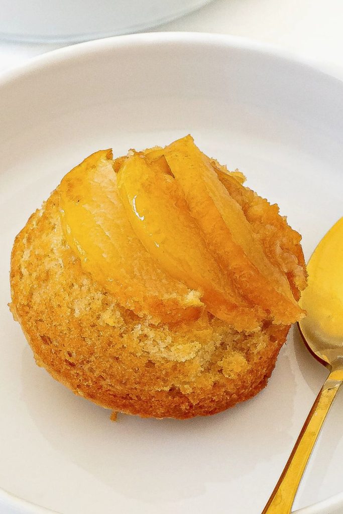 A single mini apple upside-down cake served in a white dish, topped with caramelized apple slices glistening with syrup. The golden cake base is moist and slightly crisp around the edges, paired with a gold spoon for an elegant presentation.