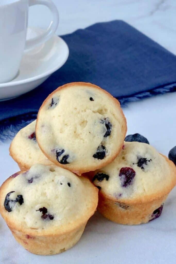 Close up of mini blueberry muffins stacked on a white surface with a soft golden exterior and tender crumb. Juicy blueberries are visible throughout each bite sized muffin, with a white coffee cup and blue napkin in the background.