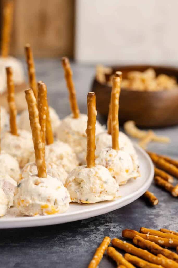 Mini cheese balls made with a creamy cheese mixture and studded with herbs and bits of bacon, each served with a pretzel stick for easy snacking, arranged neatly on a white plate with extra pretzels scattered around.