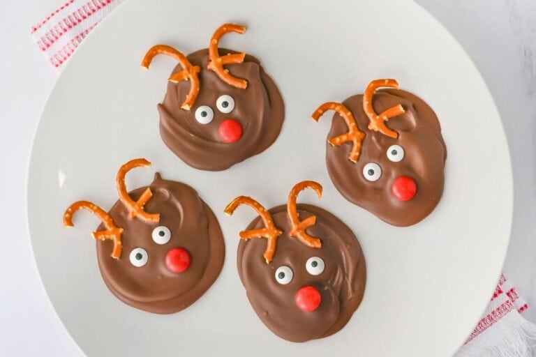 Four round chocolate bark treats decorated to look like reindeer, with mini pretzel twists for antlers, candy eyes, and red candy noses, arranged on a white plate. The playful holiday treats sit atop a white surface with a red-striped cloth peeking from underneath.