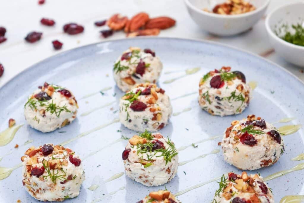 Cranberry pecan goat cheese balls studded with dried cranberries and chopped pecans and garnished with fresh dill. The bite sized appetizers are arranged on a pale blue serving platter with a golden drizzle, while extra cranberries, pecans, and small bowls of toppings sit softly out of focus in the background.