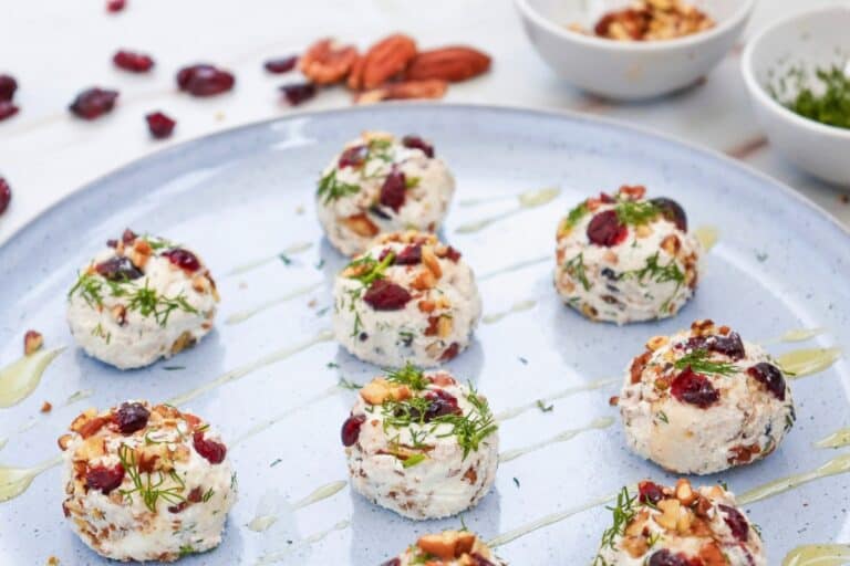 Cranberry pecan goat cheese balls studded with dried cranberries and chopped pecans and garnished with fresh dill. The bite sized appetizers are arranged on a pale blue serving platter with a golden drizzle, while extra cranberries, pecans, and small bowls of toppings sit softly out of focus in the background.