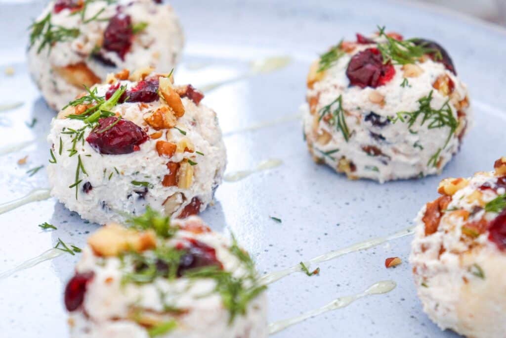 Mini cranberry pecan goat cheese balls are arranged across a pale blue platter in a close up view. The creamy white cheese balls are studded with dark red dried cranberries and chopped pecans and topped with fresh green dill while thin lines of golden honey are drizzled between them.