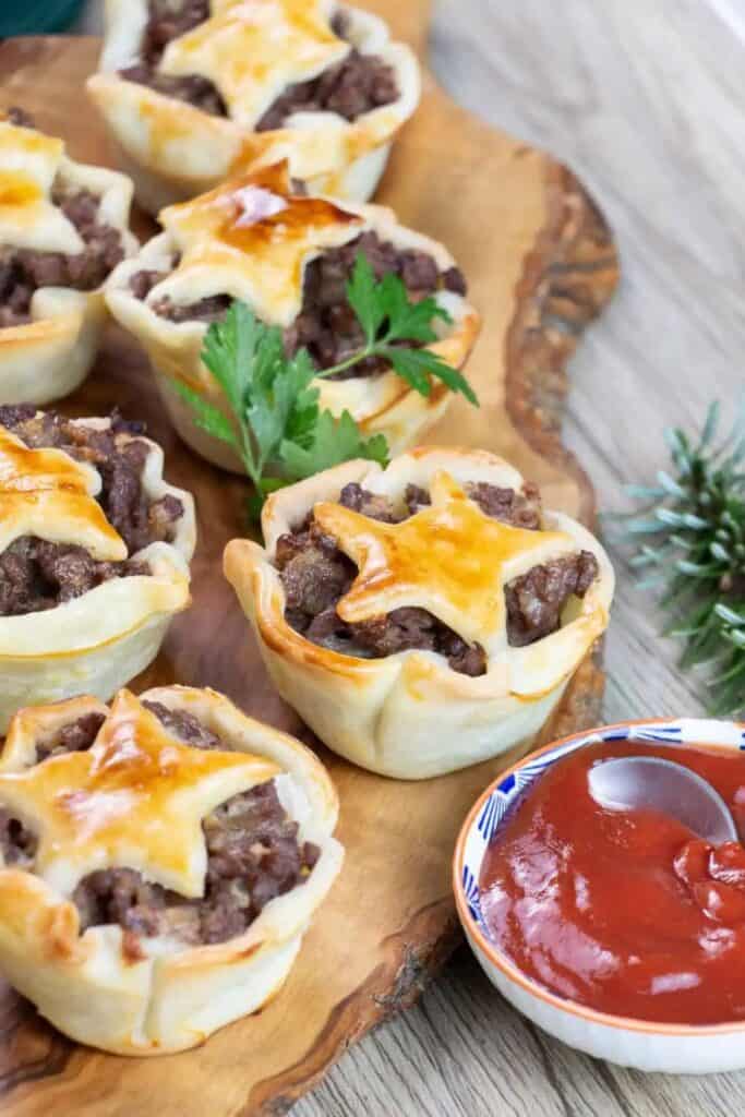 Mini Christmas meat pies filled with seasoned ground meat and topped with golden star shaped pastry crusts. The individual pies are arranged on a wooden serving board beside a small bowl of red dipping sauce.