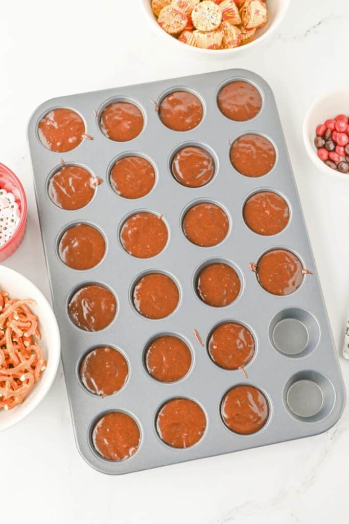 Mini muffin tin filled with raw brownie batter, ready to bake, with bowls of candy eyes, Reese’s mini cups, M&M’s, and pretzels arranged around it. The setup is part of a step-by-step process for making reindeer brownie bites.