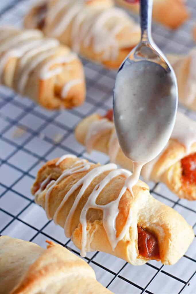 Spoon drizzling icing over mini pumpkin croissants on a metal cooling rack. 