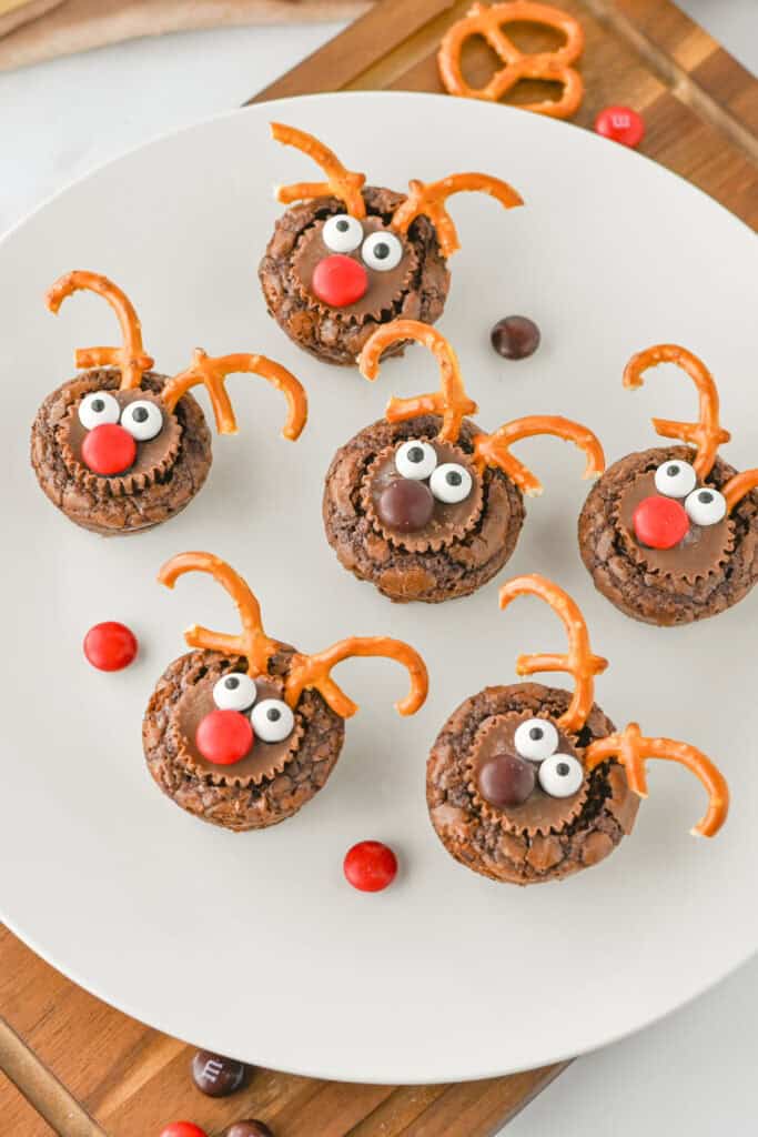 A white plate filled with Rudolph brownie bites decorated with chocolate peanut butter cups, candy eyes, red and brown candy noses, and curved pretzel pieces shaped like reindeer antlers.