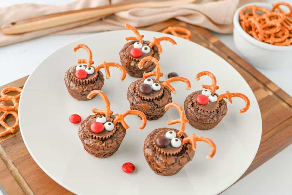 Side view of mini reindeer themed Christmas brownies on a white plate, each decorated with a Reese&rsquo;s mini cup, pretzel antlers, candy eyes, and red or brown M&M noses. The festive treats are surrounded by extra pretzels and candies.