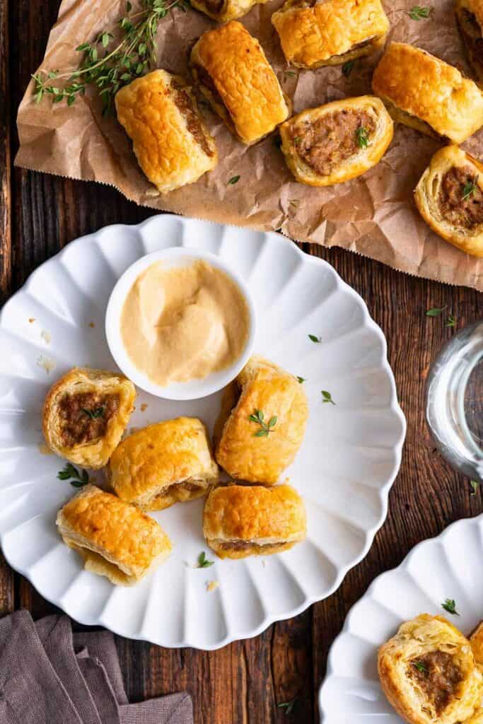 Golden puff pastry sausage rolls served on white plates with a creamy dipping sauce and fresh thyme. These bite sized Christmas Eve party appetizers are arranged on a rustic wooden table.