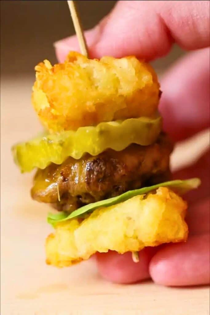 A close-up of a mini burger appetizer held together with a toothpick, featuring crispy tater tots as buns, a slice of pickle, melted cheese over a mini beef patty, and a piece of lettuce, perfectly bite-sized party snack.