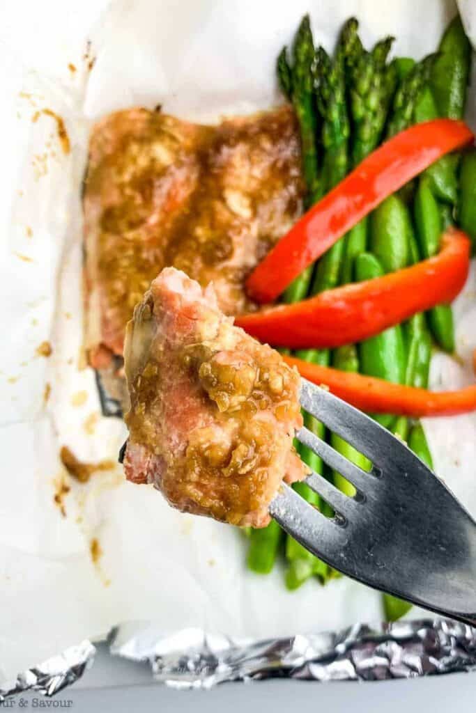 A close up of a forl with a piece of miso glazed baked salmon over a foil packet with the rest of the salmon and asparagus. 