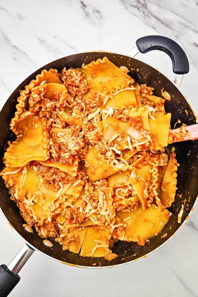Cooked lasagna noodles tossed with Italian sausage meat sauce and shredded cheese in a large skillet. The mixture is ready to be spread onto a sheet pan for the lasagna.
