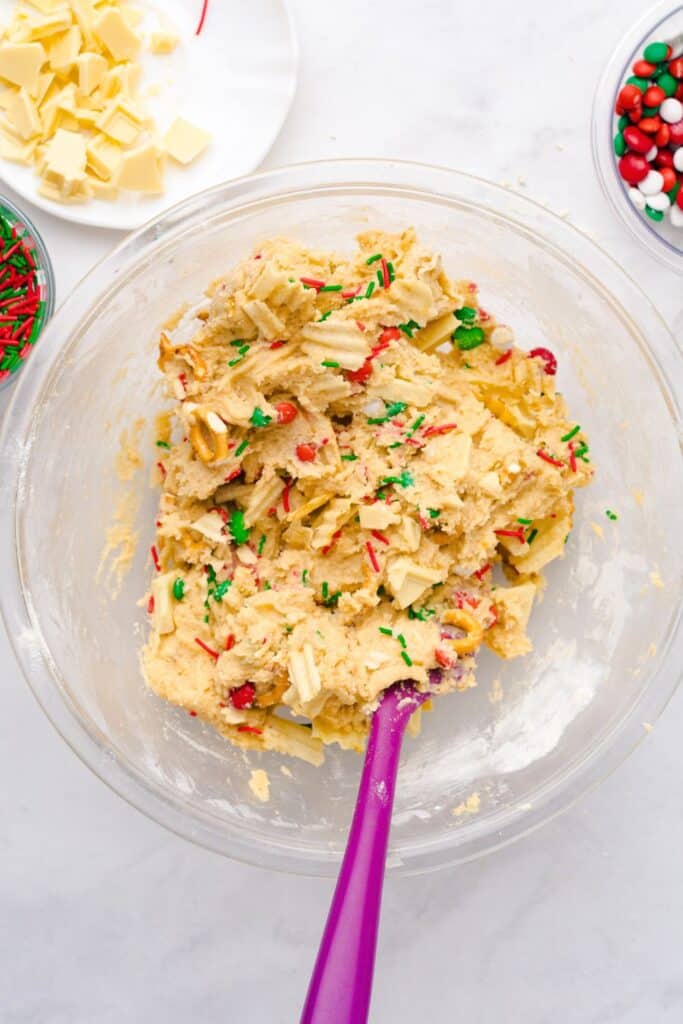 Fully mixed cookie dough in a glass bowl with festive mix-ins like red and green sprinkles, M&Ms, white chocolate chunks, pretzel pieces, and potato chips, stirred together with a purple spatula.