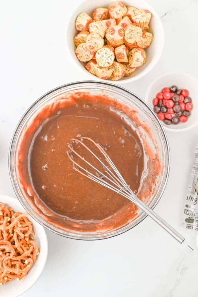 Glass bowl filled with brownie batter being mixed with a metal whisk, surrounded by bowls of unwrapped Reese’s mini cups, red and brown M&M’s, and mini pretzels on a white surface. Ingredients are prepared for making reindeer-themed holiday treats.