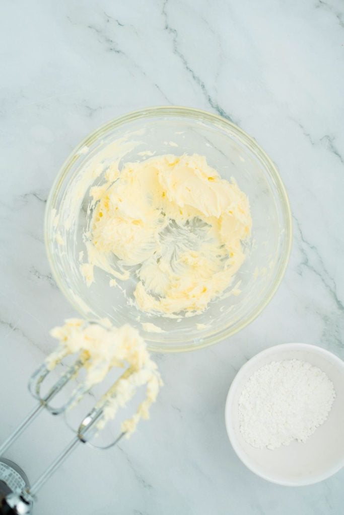 Glass bowl with creamed butter and cream cheese mixture on a marble countertop, with an electric mixer in the foreground and a small bowl of powdered sugar beside it, ready for the next step in dessert preparation.