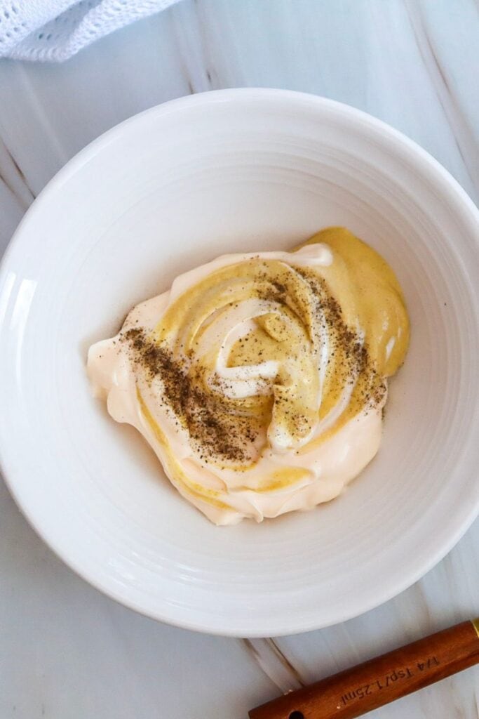 An overhead view of a ceramic bowl filled with mayo, dijon mustard, salt and pepper for potato salad. 