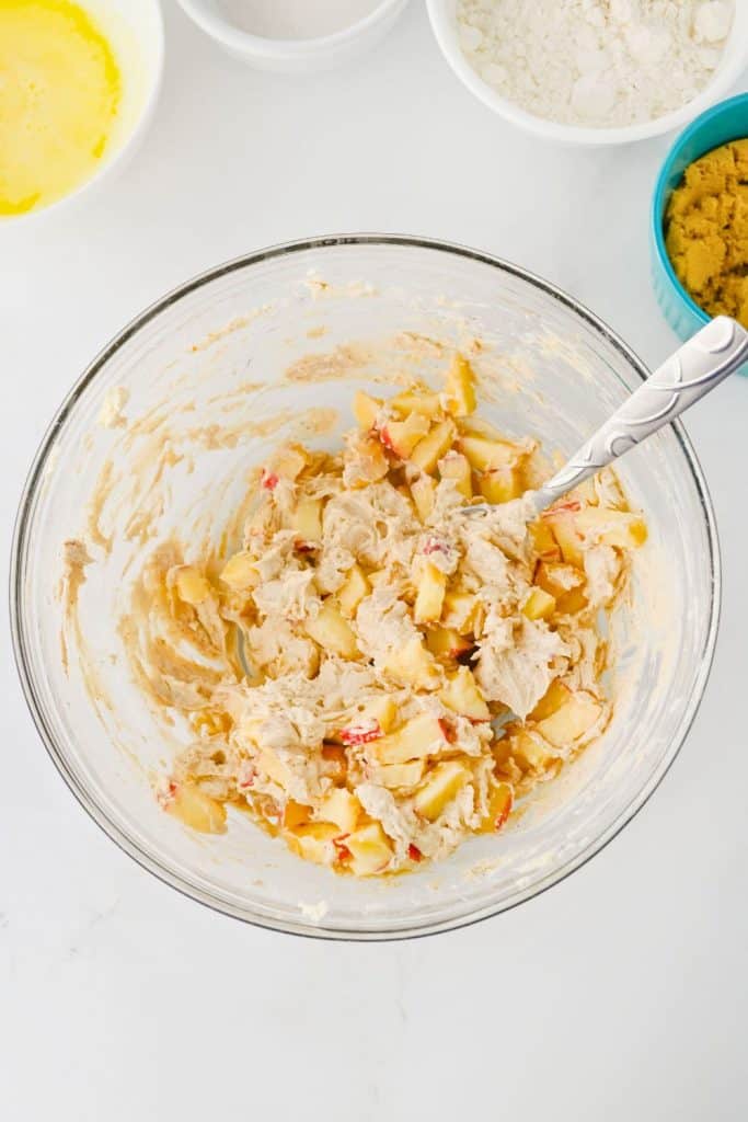 A glass bowl filled with a creamy mixture of chopped fresh apples and spiced cream cheese, being stirred with a metal spoon. Surrounding the bowl are ingredients like melted butter, sugar, flour, and brown sugar, set on a white surface.
