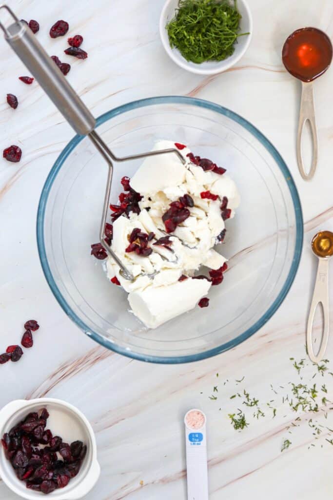 Process shot showing goat cheese and dried cranberries being mashed together in a clear glass bowl with a metal potato masher. Bowls of fresh dill and dried cranberries sit nearby with measuring spoons filled with honey, vanilla, and salt.