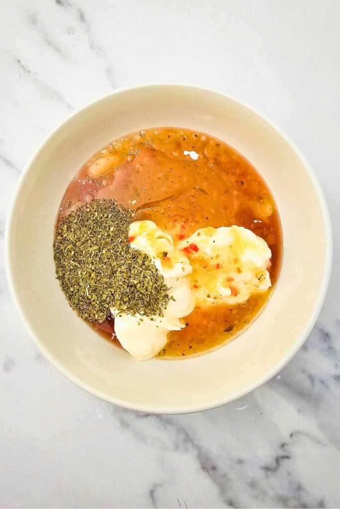 A white bowl filled with ingredients for a creamy hoagie pasta salad dressing, including mayonnaise, Italian dressing, red wine vinegar, and a heap of dried Italian seasoning. The ingredients are unmixed and sit on a light marble countertop, ready to be blended into a flavorful sauce.