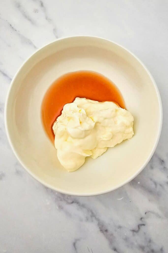 A white bowl containing a dollop of mayonnaise and a pour of red wine vinegar, ready to be mixed into a creamy dressing. The bowl sits on a light marble surface, showing a simple step in preparing Italian hoagie salad dressing.