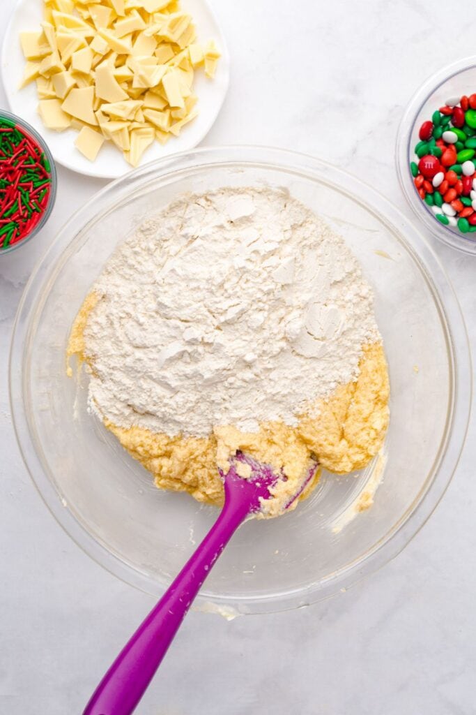 Glass bowl filled with all-purpose flour and baking soda, ready to be mixed into cookie dough, with chopped white chocolate nearby and part of a mixing bowl and whisk visible on the side.