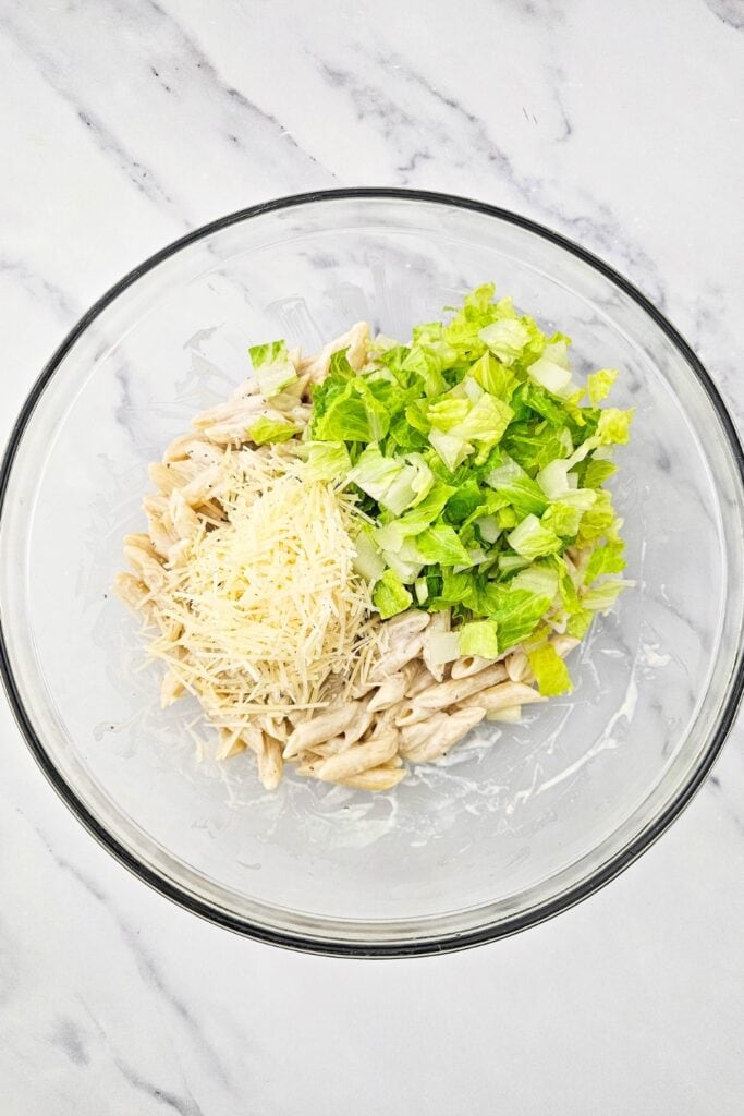 Cooked penne pasta mixed with Caesar dressing in a glass bowl topped with chopped romaine lettuce and shredded Parmesan, showing a quick family dinners recipe coming together before adding croutons.