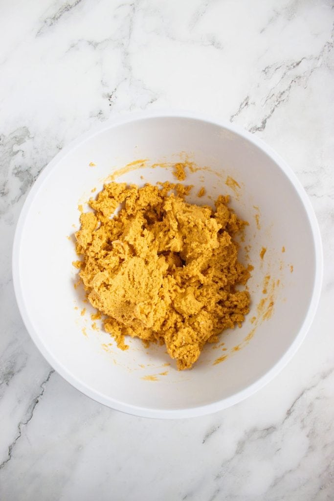 Thick, fully combined pumpkin spice cookie dough in a white mixing bowl, resting on a marble countertop. The dough has a rich orange color and a soft, slightly sticky texture, ready to chill or roll.