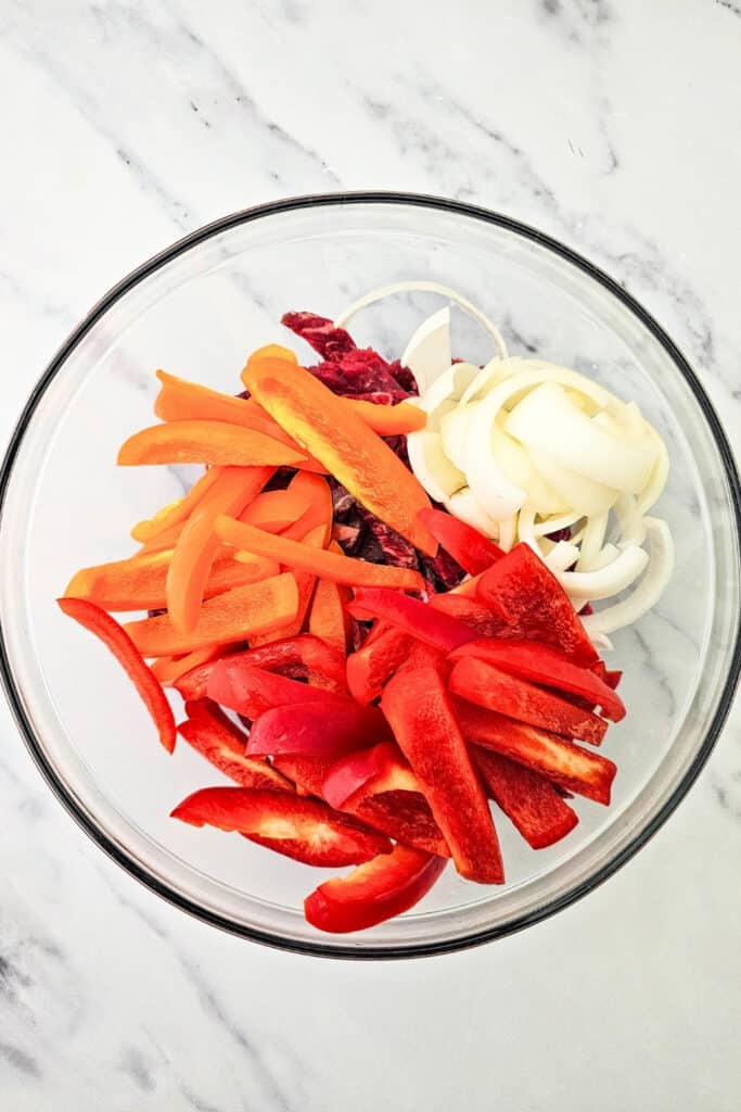 Red and orange bell pepper strips and sliced white onion are added to raw steak in a large glass mixing bowl. The sliced vegetables cover most of the steak as the fajita ingredients are prepared for mixing.