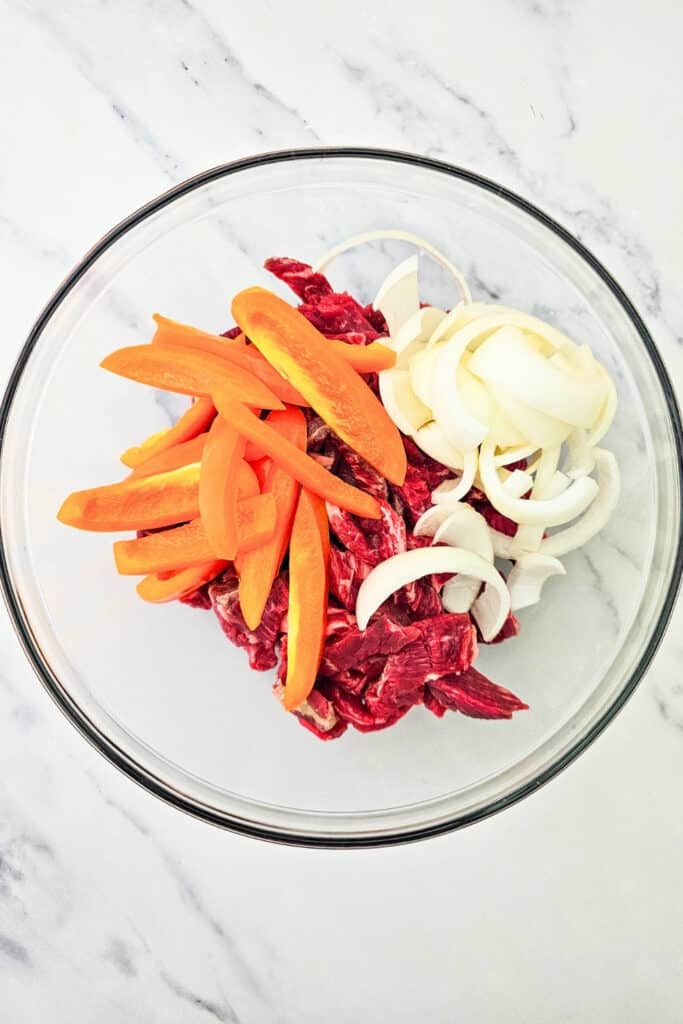 Overhead view of sliced raw steak in a glass bowl with piles of orange bell pepper strips and sliced white onion. 