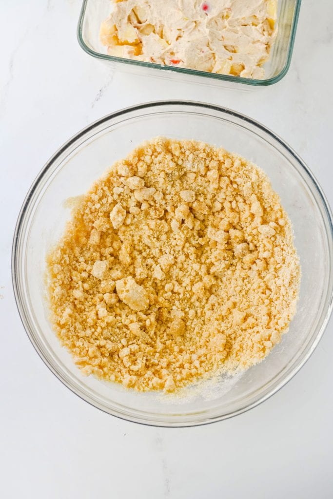 A glass mixing bowl filled with golden, crumbly streusel topping made from butter, sugar, and flour. In the background, a baking dish holds a layer of apple cream cheese dip, ready for the topping.