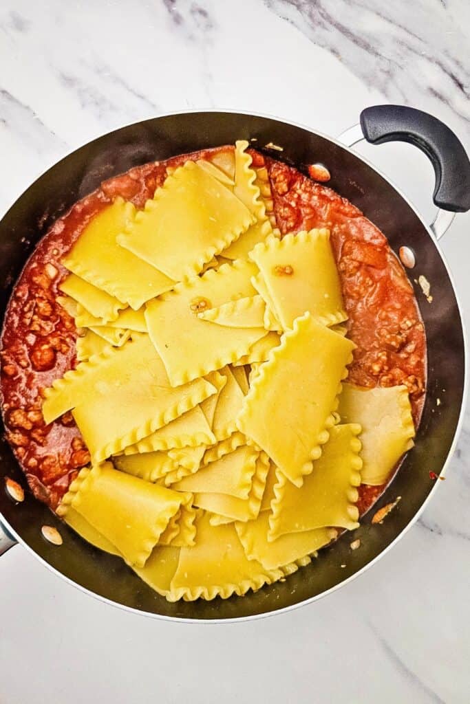 Overhead view of cooked lasagna noodles added to a skillet of Italian sausage and tomato meat sauce. The broken noodles are ready to be mixed with the sauce.