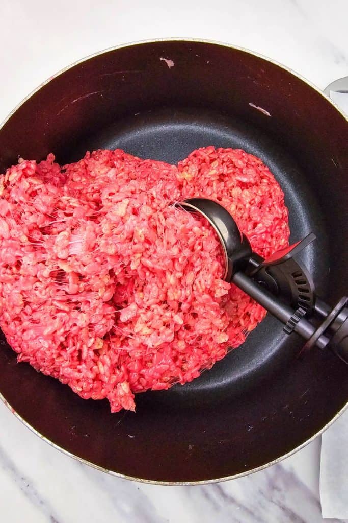 Bright red rice krispie mixture in a saucepan, being scooped with a black ice cream scoop to shape Halloween treats.