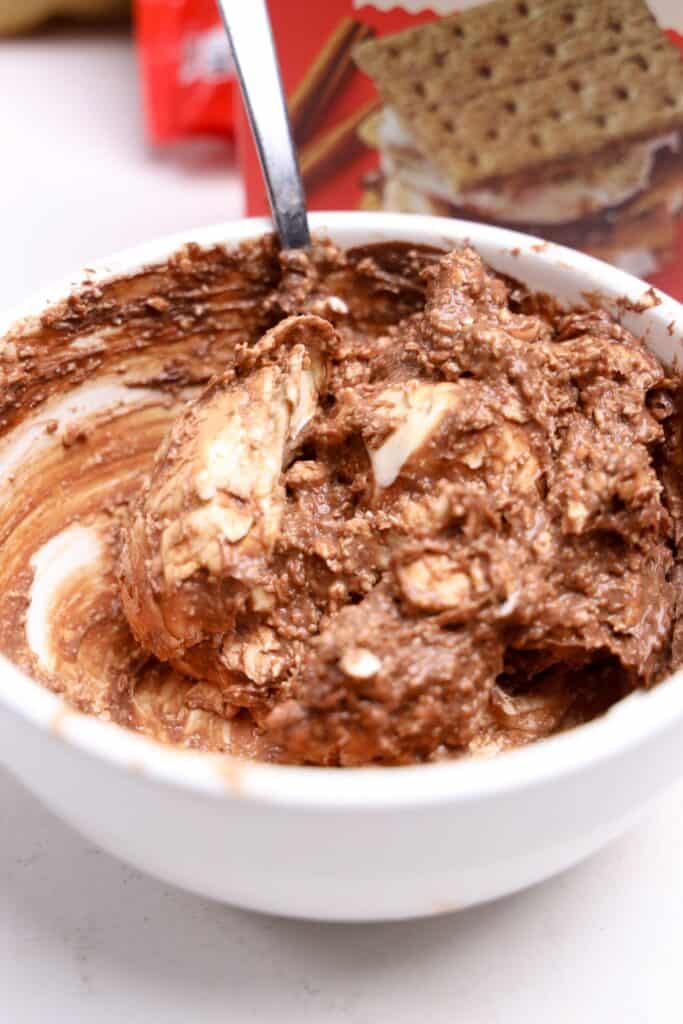 Close-up of a thick, swirled cream cheese dip being mixed in a white bowl, with visible chunks of cream cheese and chocolate spread blended together. A red graham cracker box is blurred in the background, hinting at serving suggestions.