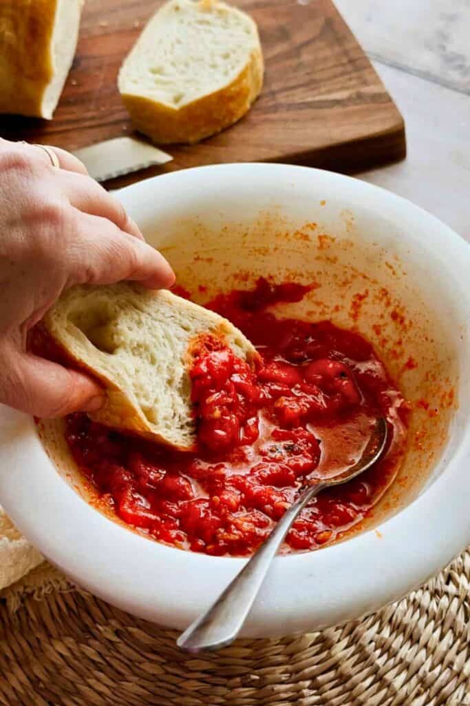 A hand dips a thick slice of crusty bread into a bowl of bright red Mojo Pico, a spicy red pepper sauce. A spoon rests in the chunky sauce while additional slices of bread sit on a wooden cutting board in the background.