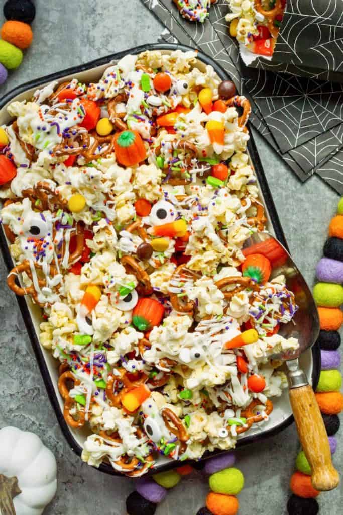 Tray of easy Halloween movie night snack mix featuring popcorn, pretzels, candy corn, candy pumpkins, candy eyes, colorful sprinkles, and a drizzle of white chocolate, all arranged festively on a metal tray with Halloween napkins and decorations in the background.