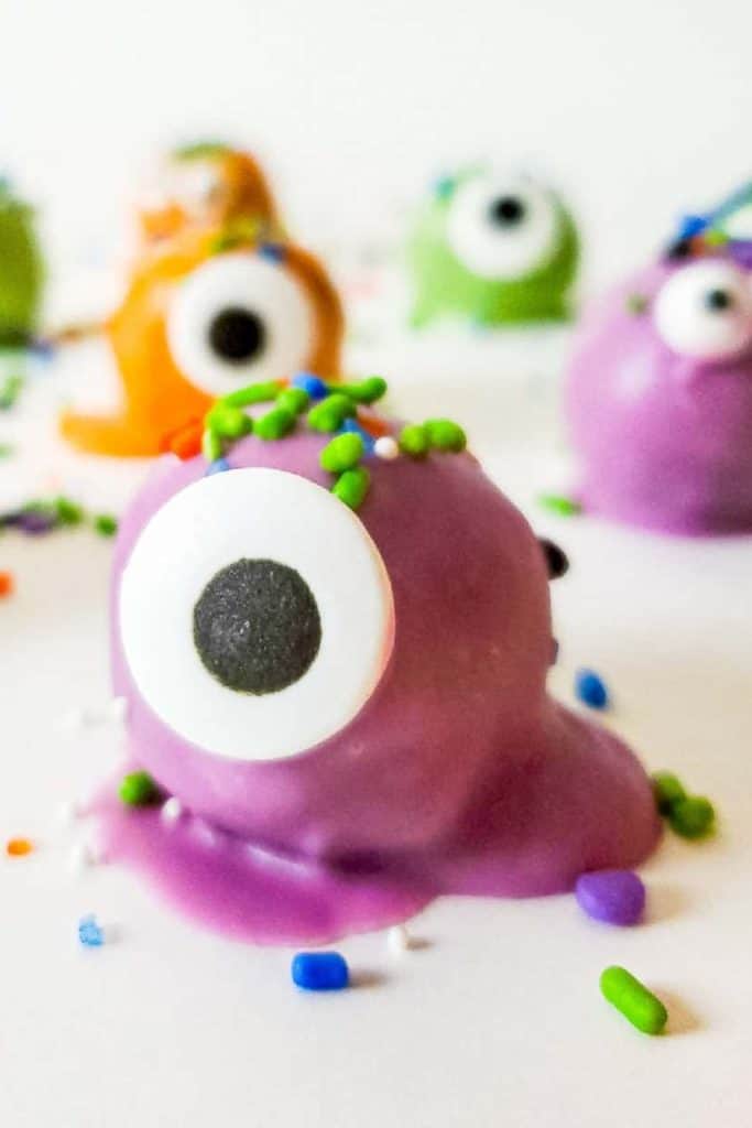 Close-up of a purple monster-themed Halloween treat covered in candy coating with a large candy eye and colorful sprinkles on top. Blurry background features more playful monster treats in green, orange, and purple, each with candy eyes and festive decorations.