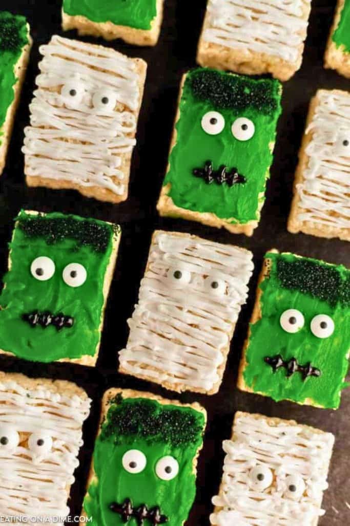Halloween Rice Krispies treats decorated as green Frankenstein faces with black sprinkle hair and stitched mouths, and white mummy faces with candy eyes and icing bandages.