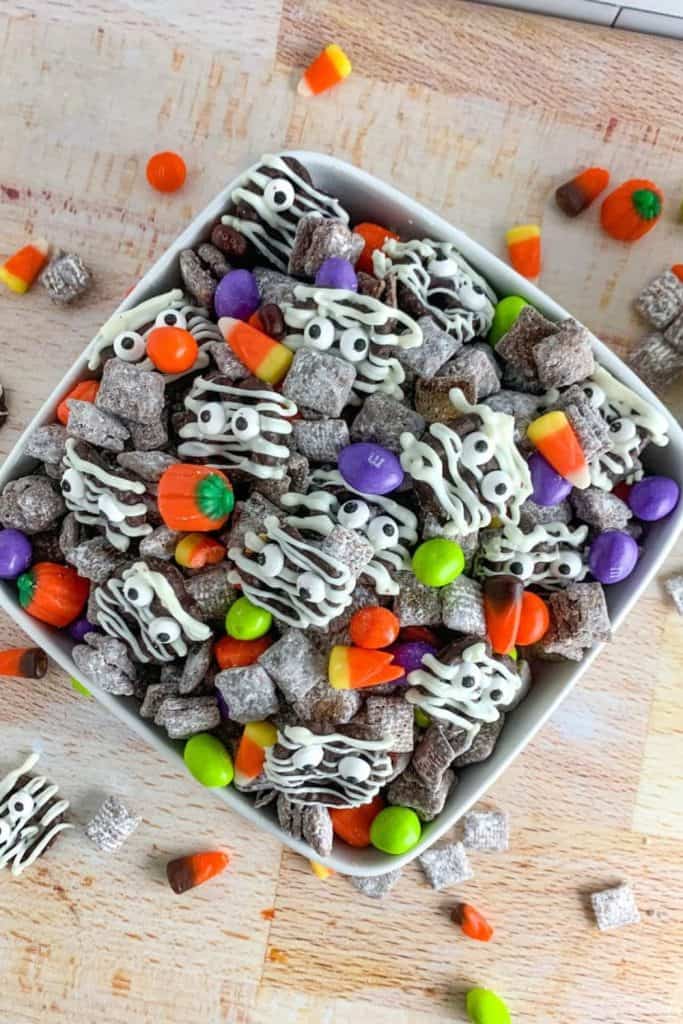 Square white bowl filled with Halloween-themed snack mix featuring chocolate-covered cereal squares drizzled with white chocolate and candy eyes to resemble mummies. Mix also includes colorful candy pieces like candy corn, pumpkins, and vibrant M&M style chocolates.