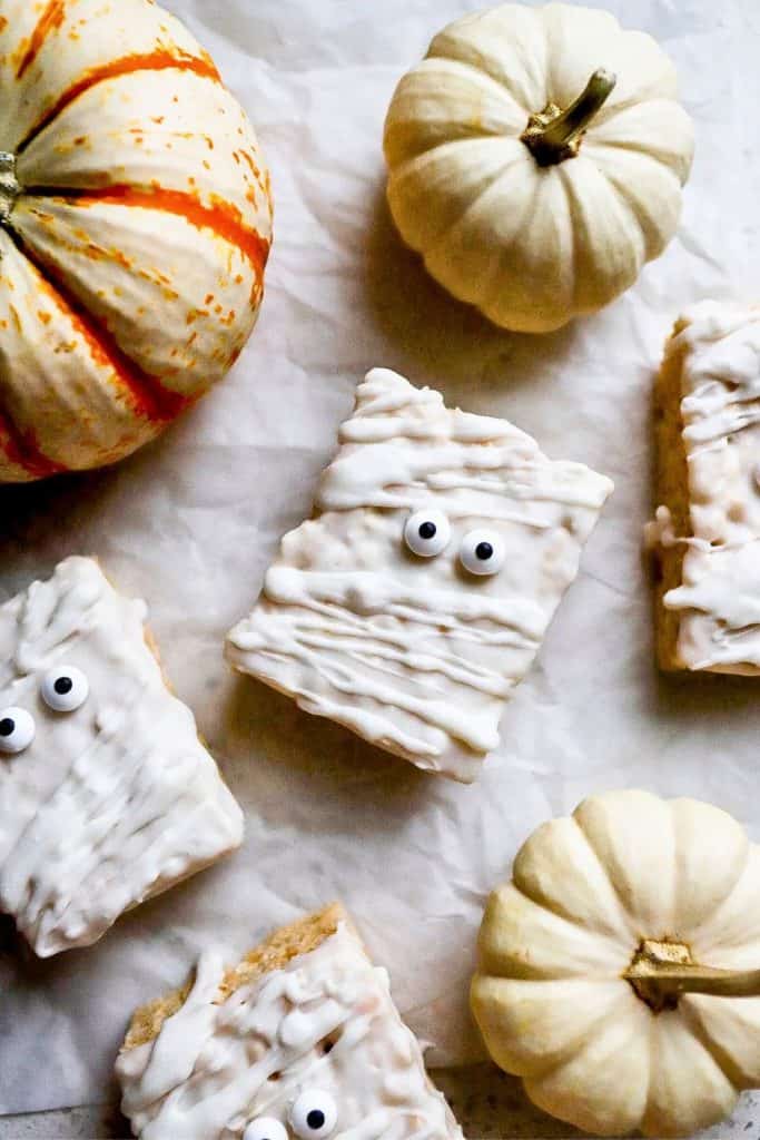 White chocolate–covered Rice Krispies treats decorated like mummies with candy eyes, arranged on parchment paper alongside small white and orange-striped pumpkins.