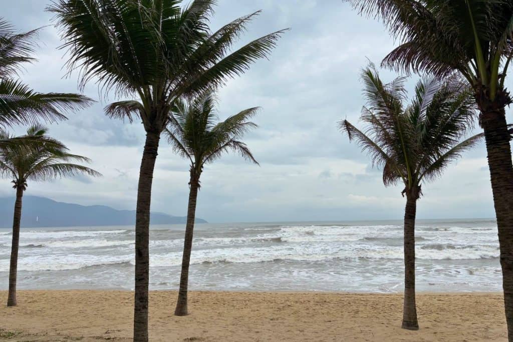 Palm trees line the sandy shore of My Khe Beach under a cloudy sky, with gentle waves rolling in from the sea. In the distance, Son Tra Peninsula rises along the coastline, and the white statue of Linh Ung Pagoda is faintly visible against the hillside.