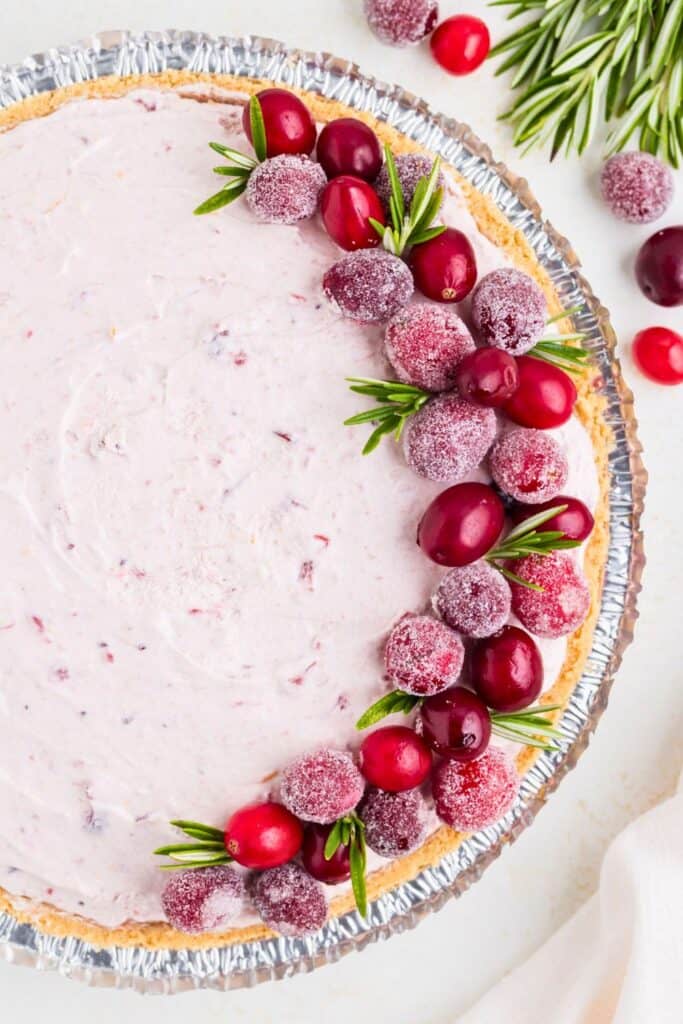No Bake Cranberry Pie with a pale pink creamy filling in a graham cracker crust. Fresh and sugar coated cranberries with sprigs of rosemary decorate one side of the pie for a festive finish.